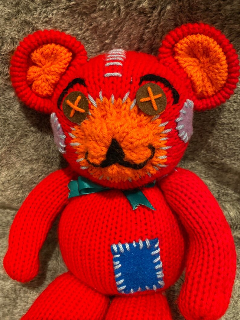 May include: A red knitted teddy bear with orange ears and a stitched-on blue patch. The bear has a stitched-on mustache and X-shaped eyes.