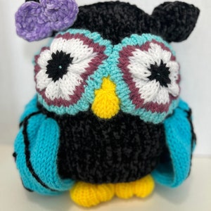 May include: A black knitted owl with blue wings and yellow feet. The owl has large white eyes with black pupils and a purple flower on its head.