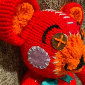 May include: A red knitted teddy bear with orange, blue, and purple patches. The bear has a brown button for an eye and a black stitched mouth.