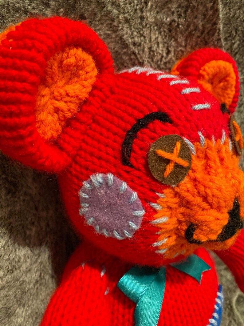 May include: A red knitted teddy bear with orange, blue, and purple patches. The bear has a brown button for an eye and a black stitched mouth.