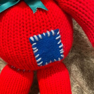 May include: A red knitted toy with a green bow and a blue patch sewn on the front. The toy has a stitched-on face and is made of a soft, plush material.
