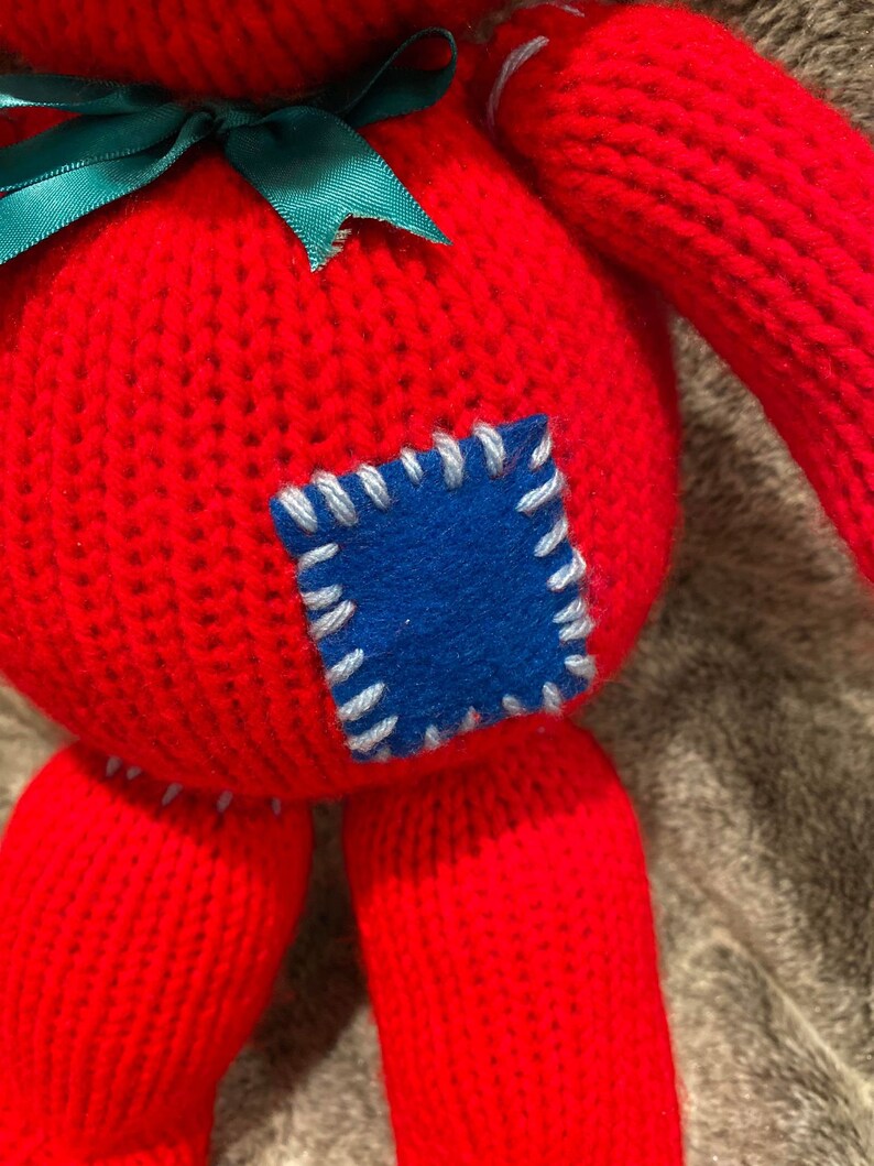 May include: A red knitted toy with a green bow and a blue patch sewn on the front. The toy has a stitched-on face and is made of a soft, plush material.
