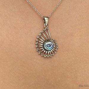 May include: A silver necklace with a shell-shaped pendant featuring an iridescent abalone inlay. The pendant is detailed with a delicate, openwork design.