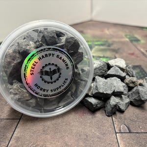 May include: A clear plastic container filled with small, dark grey rocks. The container has a black and white label that reads "Steel Harpy Gaming Hobby Supplies".