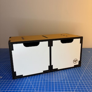 May include: A small, two-drawer storage box with a white interior and black trim. The box is made of cardboard and has a simple, modern design.