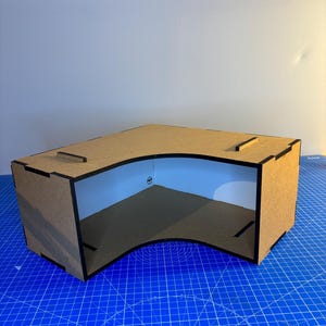 May include: A cardboard corner shelf with a curved front edge. The shelf is brown with black edges and is sitting on a blue cutting mat.