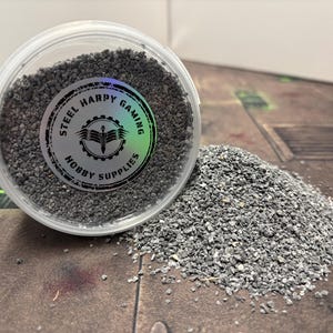 May include: A clear plastic container filled with grey gravel, spilling out onto a wooden surface. The container has a black and white label that reads "Steel Harpy Gaming Hobby Supplies".