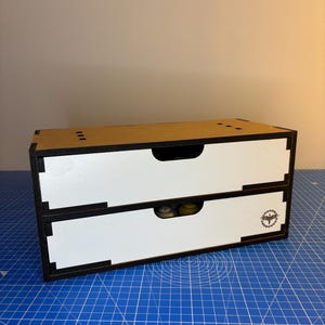 May include: A two-drawer wooden storage box with a white interior and black trim. The box has a brown top and a logo on the front of the bottom drawer.