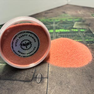 Op de afbeelding: Een klein bakje met roze zand met een label met de tekst "Steel Harpy Gaming Hobby Supplies".