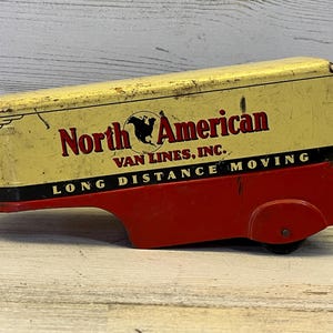 May include: A vintage metal toy trailer with a cream-colored top and a red base. The side features the text North American VAN LINES, INC. and LONG DISTANCE MOVING in black and red lettering, along with a black map graphic of North America.