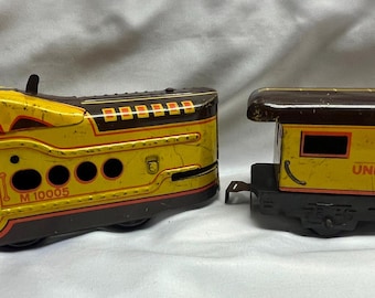 May include: A vintage tin toy train set featuring a yellow and brown locomotive marked M 10005 and a matching yellow and brown caboose labeled Union Pacific 3824. Both train cars have painted details, rectangular windows, and black metal wheels.