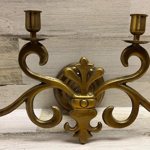 May include: A wall-mounted brass candelabra featuring four candle holders arranged on curved, scrolling arms. The central section displays a decorative fleur-de-lis motif, and the entire piece has a polished metallic finish with a warm golden tone.