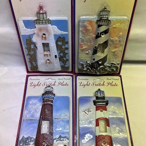 May include: Four hand-painted decorative light switch plates in original packaging. Each plate features a three-dimensional lighthouse design with unique color schemes, including white, black, red, and brown, set against painted coastal scenes.
