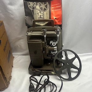 May include: A vintage Bell & Howell Monterey 8mm projector in a dark olive-gray finish, featuring a top-mounted handle, a lens assembly, and a separate black film reel. It includes an instruction manual titled How to use your Bell & Howell MONTEREY 8mm PROJECTOR and a power cord.
