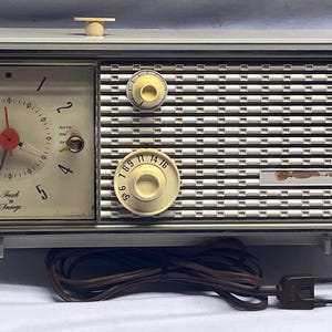 May include: A rectangular off-white electric alarm clock radio featuring an analog clock face on the left with a red second hand and the text Touch 'n Snooze. The right side has a white speaker grille with two circular knobs for tuning and volume.