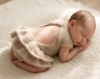 Könnte beinhalten: Ein beigefarbener Strickstrampler mit tiefem Rückenausschnitt und zwei Lagen Rüschen am unteren Teil. Das Kleidungsstück besteht aus weichem, strukturiertem Garn und wird von einem schlafenden Säugling getragen, der auf einer beigen Decke liegt.