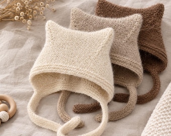 Peut inclure: Trois bonnets tricotés avec des détails en forme d'oreilles de chat pointues, chacun doté de longs liens pour le menton. Les bonnets sont disposés en rangée, dans des tons crème, beige clair et brun foncé, avec un point jersey simple.