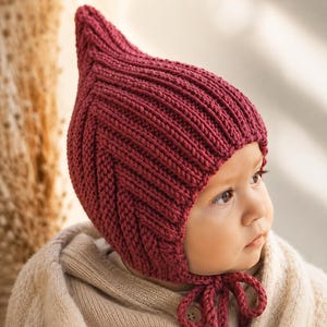 May include: A burgundy knit baby bonnet with a pointed top and ribbed texture. The bonnet has ties under the chin and is worn by a baby. The baby is wearing a beige sweater.