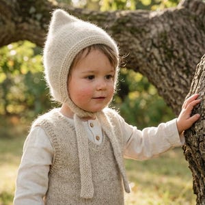 Könnte beinhalten: Eine cremefarbene Strickhaube mit spitzer Oberseite und Kinnbändern, getragen über einer passenden cremefarbenen Strickweste, die über einem langärmeligen Hemd mit kleinen Knöpfen am Ausschnitt liegt, aus weichem, strukturiertem Garn.