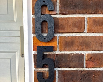 May include: Black metal house numbers "650" mounted on a red brick wall. The numbers are in a modern font and have a textured finish. The brick wall has a white trim and a white garage door in the background.
