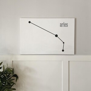 May include: A white canvas print with a black line drawing of the constellation Aries. The word "aries" is printed in black letters below the constellation.