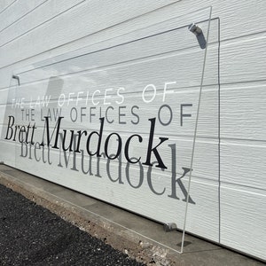 Pu&ograve; includere: Un'insegna trasparente in acrilico con la scritta "The Law Offices of Brett Murdock" &egrave; fissata a una parete bianca. Il testo &egrave; in nero, bianco e grigio, con il nome in un carattere elegante e in grassetto.
