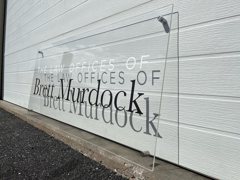 Pu&ograve; includere: Un'insegna trasparente in acrilico con la scritta "The Law Offices of Brett Murdock" &egrave; fissata a una parete bianca. Il testo &egrave; in nero, bianco e grigio, con il nome in un carattere elegante e in grassetto.