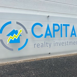 Pu&ograve; includere: Un'insegna in vetro smerigliato con le parole "CAPITAL" in blu e "realty investments" in grigio. L'insegna presenta un logo con una casa blu, una freccia gialla e verde e un design circolare blu e grigio. L'insegna &egrave; montata su un muro bianco.