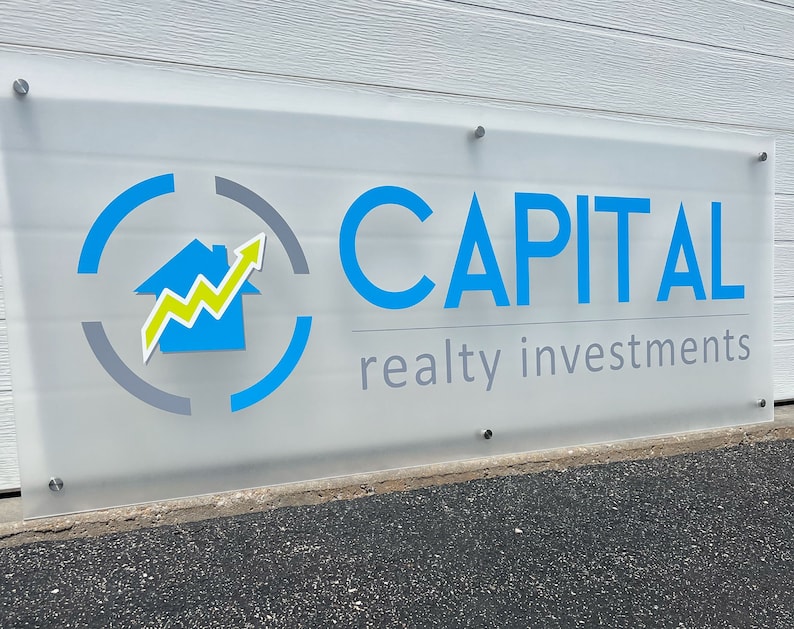 Pu&ograve; includere: Un'insegna in vetro smerigliato con le parole "CAPITAL" in blu e "realty investments" in grigio. L'insegna presenta un logo con una casa blu, una freccia gialla e verde e un design circolare blu e grigio. L'insegna &egrave; montata su un muro bianco.