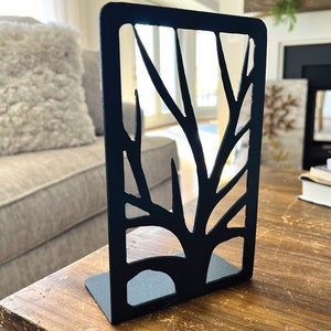 May include: Black metal bookends shaped like a tree with a cutout design. The bookends are on a wooden table.