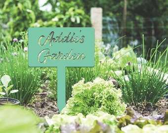 Può includere: Un cartello da giardino turchese con le parole "Addie's Garden" ritagliate. È posizionato in un'aiuola con piante verdi e lattuga. Lo sfondo è sfocato con più verde.