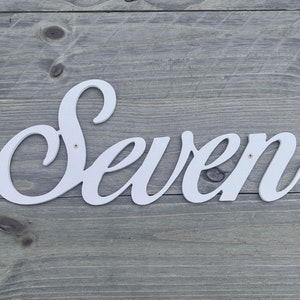 Può includere: Un cartello di legno bianco con la parola "Seven" in un font corsivo.