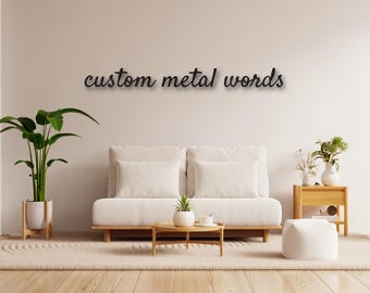 May include: A white couch with two pillows sits in a living room with a wooden floor. A potted plant is on the left side of the couch and a small wooden table with a potted plant and a cup sits in front of the couch. The wall behind the couch is white with the text "custom metal words" in black cursive writing.