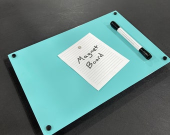 Puede incluir: Una pizarra magnética rectangular de color azul claro con esquinas redondeadas. Un papel rayado con las palabras "Magnet Board" está sujeto con un imán. Un marcador negro descansa sobre la pizarra. Sujetadores negros en cada esquina.