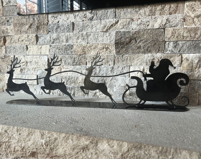 Puede incluir: Silueta de metal negro de Pap&aacute; Noel en un trineo tirado por tres renos. Los renos est&aacute;n en posici&oacute;n de carrera con las astas levantadas. El trineo tiene un respaldo curvo y un solo pat&iacute;n. La silueta est&aacute; sobre una base de metal negro.