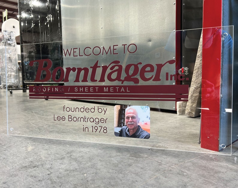 Pu&ograve; includere: Segno in acrilico trasparente con testo bordeaux: "WELCOME TO Borntrager Inc. ROOFING / SHEET METAL". Include una foto di Lee Borntrager e il testo "fondata da Lee Borntrager nel 1978".