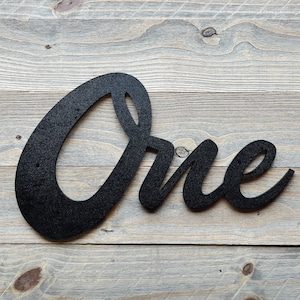 Könnte beinhalten: Schwarzes Metallschild mit dem Wort "One" in einer kursiven Schriftart. Das Schild ist auf einer verwitterten, grauen Holzoberfläche montiert. Das Schild ist ein dekorativer Artikel.