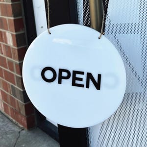 Op de afbeelding: Een rond wit bord met zwarte tekst dat "OPEN" zegt, hangend aan een touwtje.
