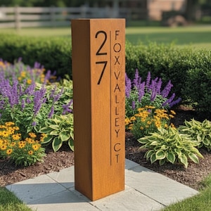 May include: A rectangular, weathered metal address sign with the number "27" in a bold, black font. The text "FOX VALLEY CT" is vertically aligned on the right side. The sign is set in a garden with colorful flowers and greenery.