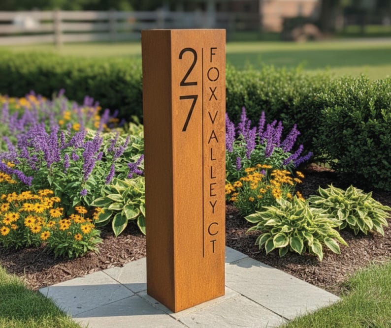May include: A rectangular, weathered metal address sign with the number "27" in a bold, black font. The text "FOX VALLEY CT" is vertically aligned on the right side. The sign is set in a garden with colorful flowers and greenery.