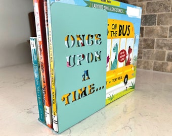 May include: A light blue metal bookend with the words "ONCE UPON A TIME..." cut out. The bookend holds several children's books, including "The Dinos on the Bus" and "How Do You Take a Bath?" The bookend is on a white surface.