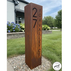 May include: A rectangular, weathered metal address sign with the number "27" and "FOX VALLEY CT" in black lettering. The sign is a rusty brown color and stands upright in a grassy yard. The sign is made by Pearl Sign Co.