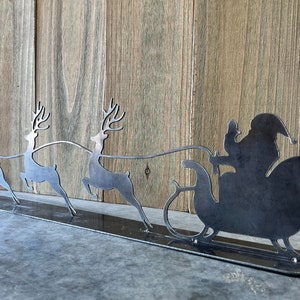 Puede incluir: Una silueta de metal de Pap&aacute; Noel montando en un trineo tirado por tres renos. Los renos est&aacute;n en fila, con Pap&aacute; Noel en la parte de atr&aacute;s. La silueta est&aacute; sobre una base de metal negro.