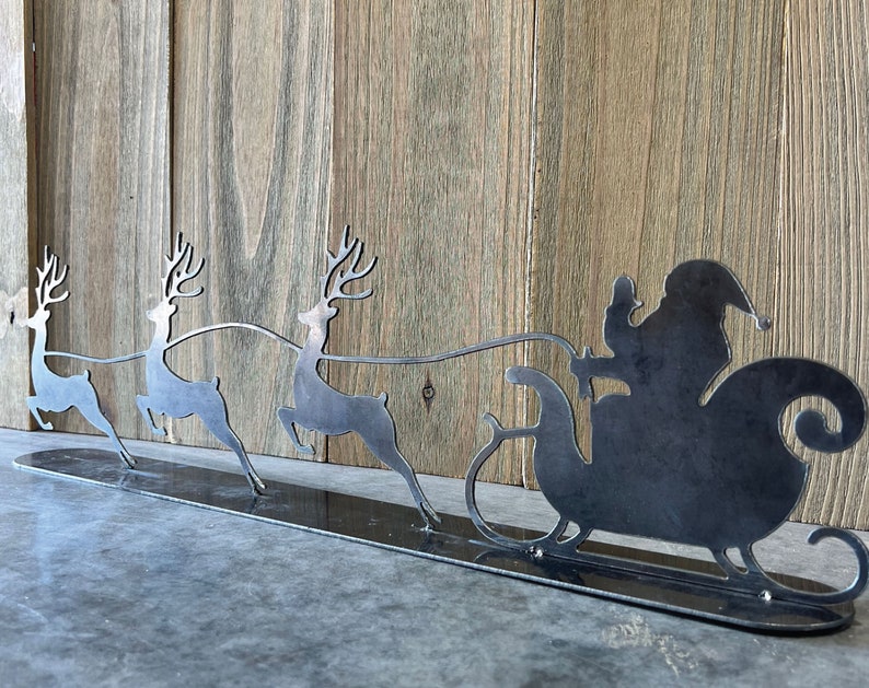 Puede incluir: Una silueta de metal de Pap&aacute; Noel montando en un trineo tirado por tres renos. Los renos est&aacute;n en fila, con Pap&aacute; Noel en la parte de atr&aacute;s. La silueta est&aacute; sobre una base de metal negro.