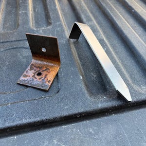 May include: A rusty L-shaped metal bracket with two circular holes and a long, silver-colored metal stake with a pointed end and a bent top section. Both metal components are positioned separately on a dark, textured surface.