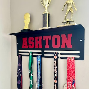 May include: Black metal medal display shelf with the name "ASHTON" in red letters. The shelf holds trophies and medals with colorful ribbons. A gold basketball trophy and soccer player trophy are on top.