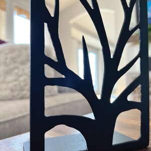 May include: Black metal bookend shaped like a tree with a cutout design. The bookend is on a wooden surface.