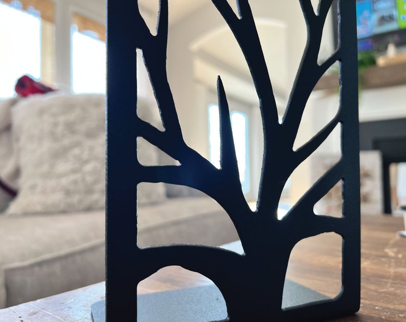 May include: Black metal bookend shaped like a tree with a cutout design. The bookend is on a wooden surface.
