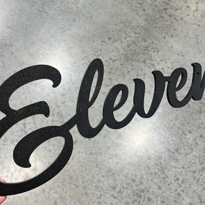 Può includere: Decorazione da parete in metallo nero con la parola "Eleven" in un font corsivo.