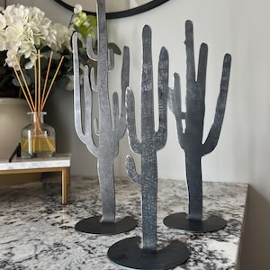 Peut inclure: Ensemble de trois sculptures de cactus en métal. Les sculptures sont en métal foncé avec une finition vieillie. Les cactus sont posés sur un comptoir gris.